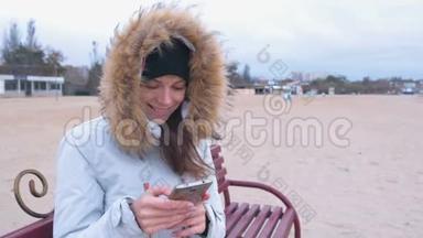 一个穿着白色羽绒服的女人坐在沙滩上的长凳上，用手机在社交媒体上打字。